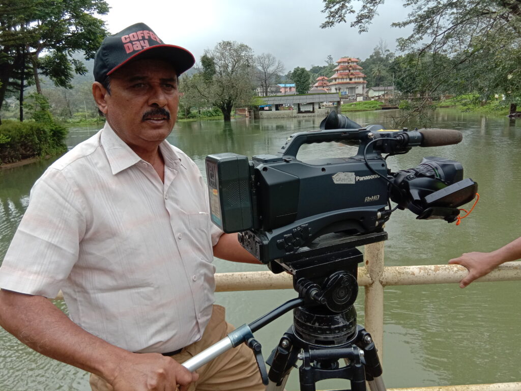 A man stands by a lake, holding a Panasonic video camera on a tripod, with greenery and a temple in the background.